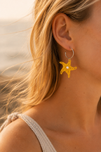 Femme portant les boucles d'oreilles Merea étoile de mer jaune en micro-macramé au bord de la mer