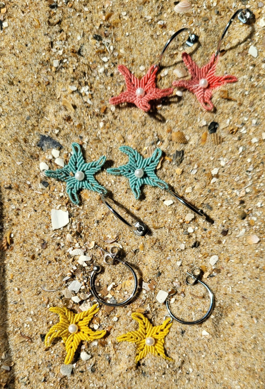 Les trois coloris des boucles d'oreilles Merea, corail, vert d'eau et jaune moutarde, posés sur le sable et des coquillages